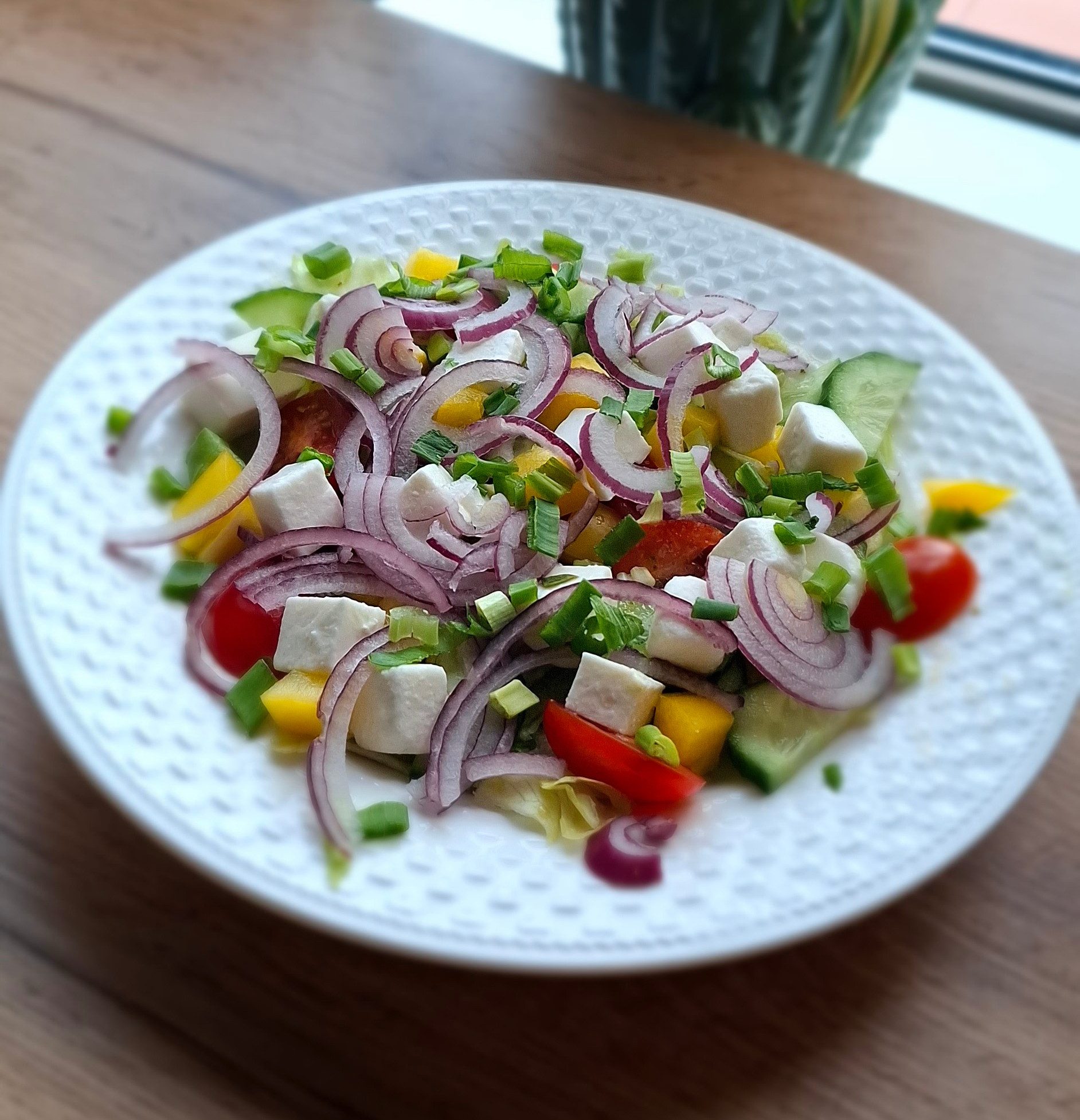 Sałatka z czerwoną cebulą, serem feta, ogórkiem, pomidorkami, papryką i szczypiorkiem na talerzu.