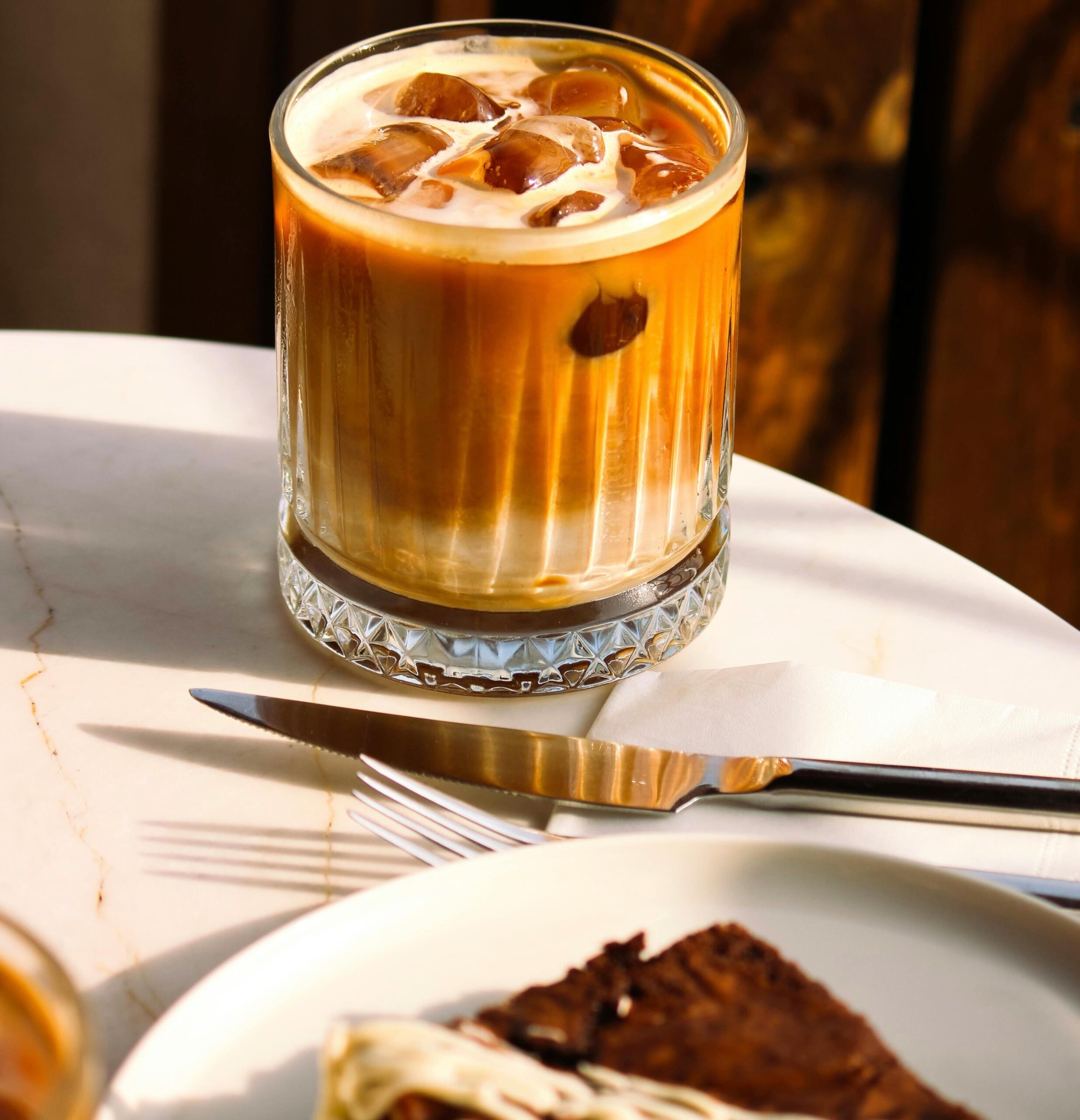 Szklanka z lodową kawą na stole obok noża, widelca i częściowo widocznego ciasta.