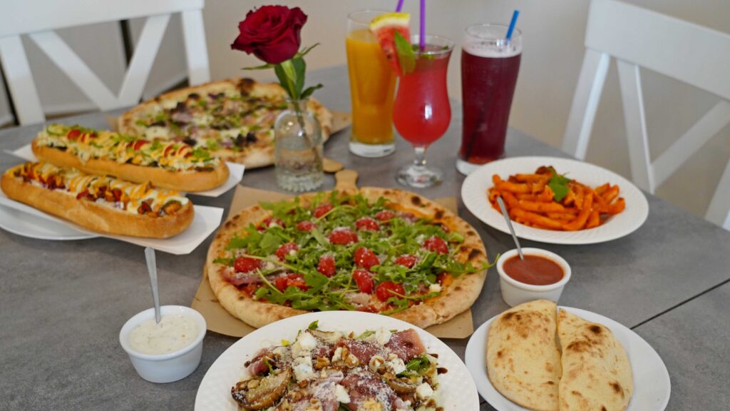 Różnorodne jedzenie na szarym stole, w tym pizza, kanapki, makaron, sosy i trzy napoje.