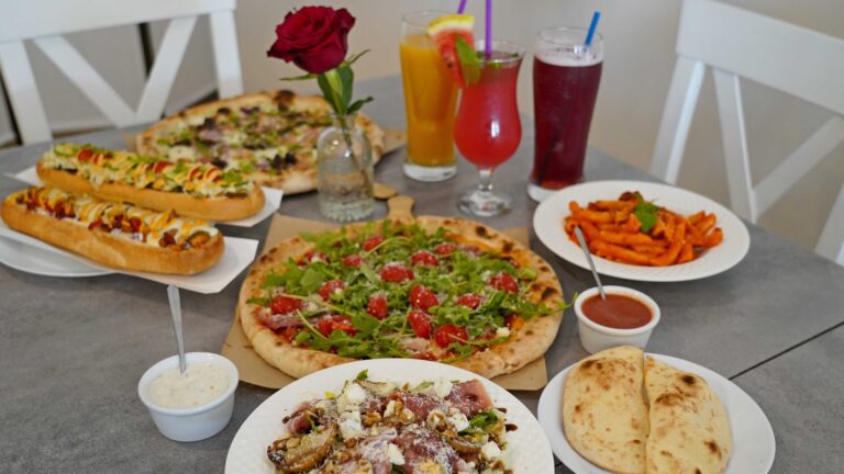 Różnorodne jedzenie na szarym stole, w tym pizza, kanapki, makaron, sosy i trzy napoje.