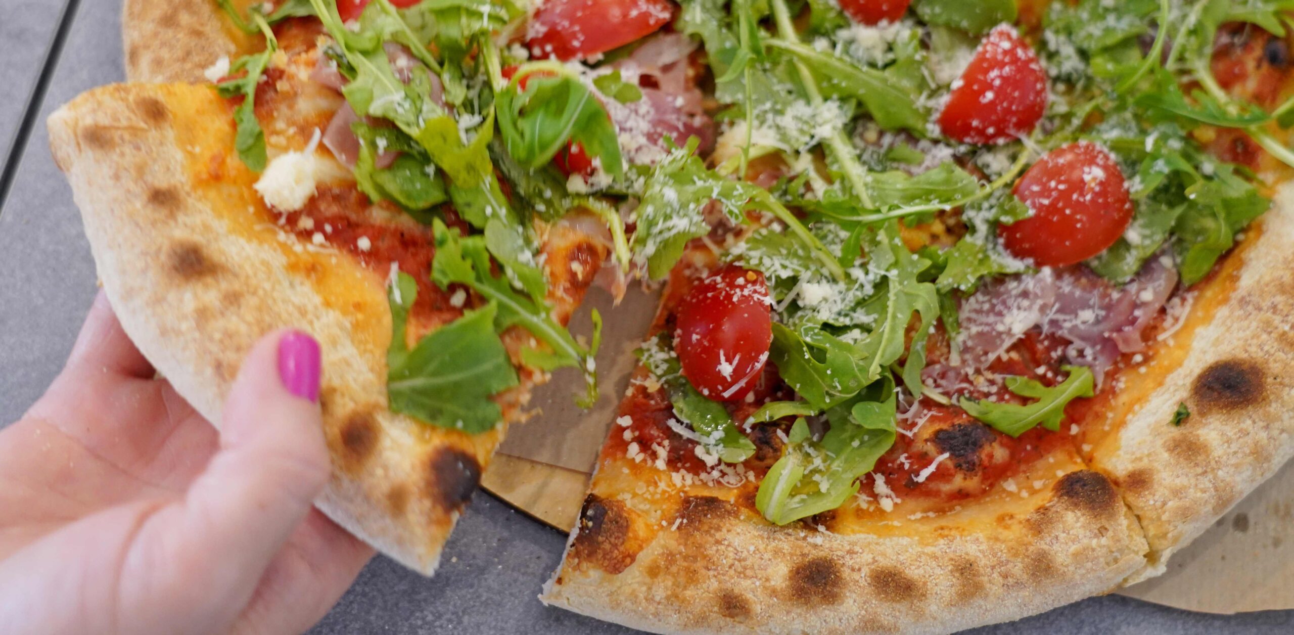 Pizza z rukolą, pomidorkami, szynką i serem, z kawałkiem trzymanym w ręku.