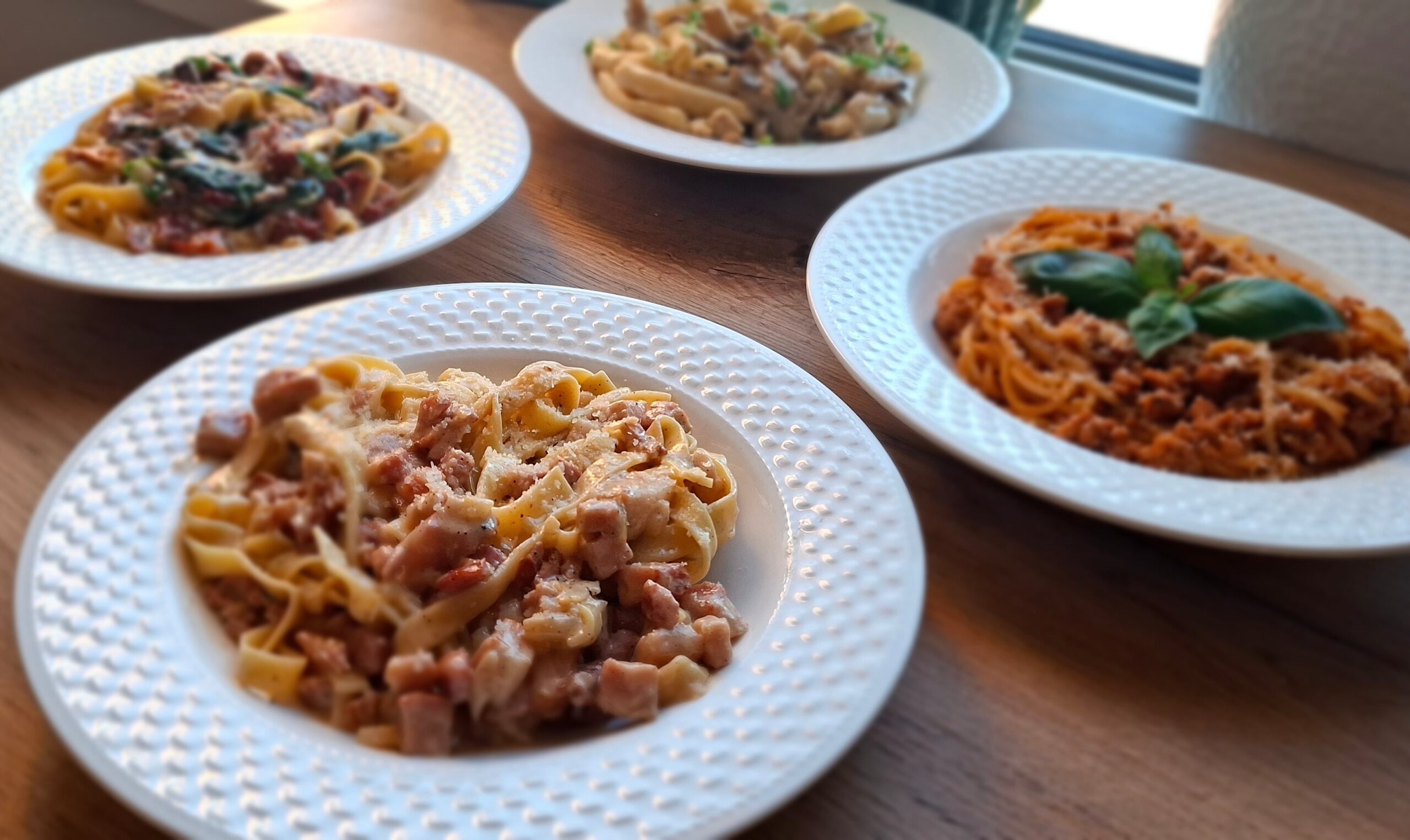 Kilka porcji różnych makaronów podanych na białych talerzach na drewnianym stole.